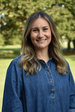A smiling woman with long, wavy hair wearing a denim shirt stands outdoors in a green park setting.