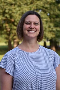 A smiling woman with short hair, wearing a striped shirt, stands outdoors with trees in the background.