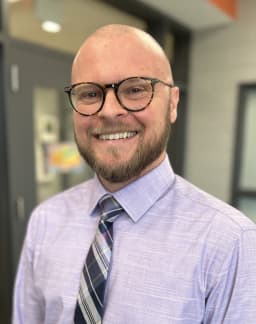 A smiling man with a bald head and glasses, wearing a light purple dress shirt and a striped tie, stands in a brightly lit indoor setting.
