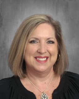 A smiling woman with shoulder-length blonde hair, wearing a black top and a necklace, poses for a professional headshot against a gray background.