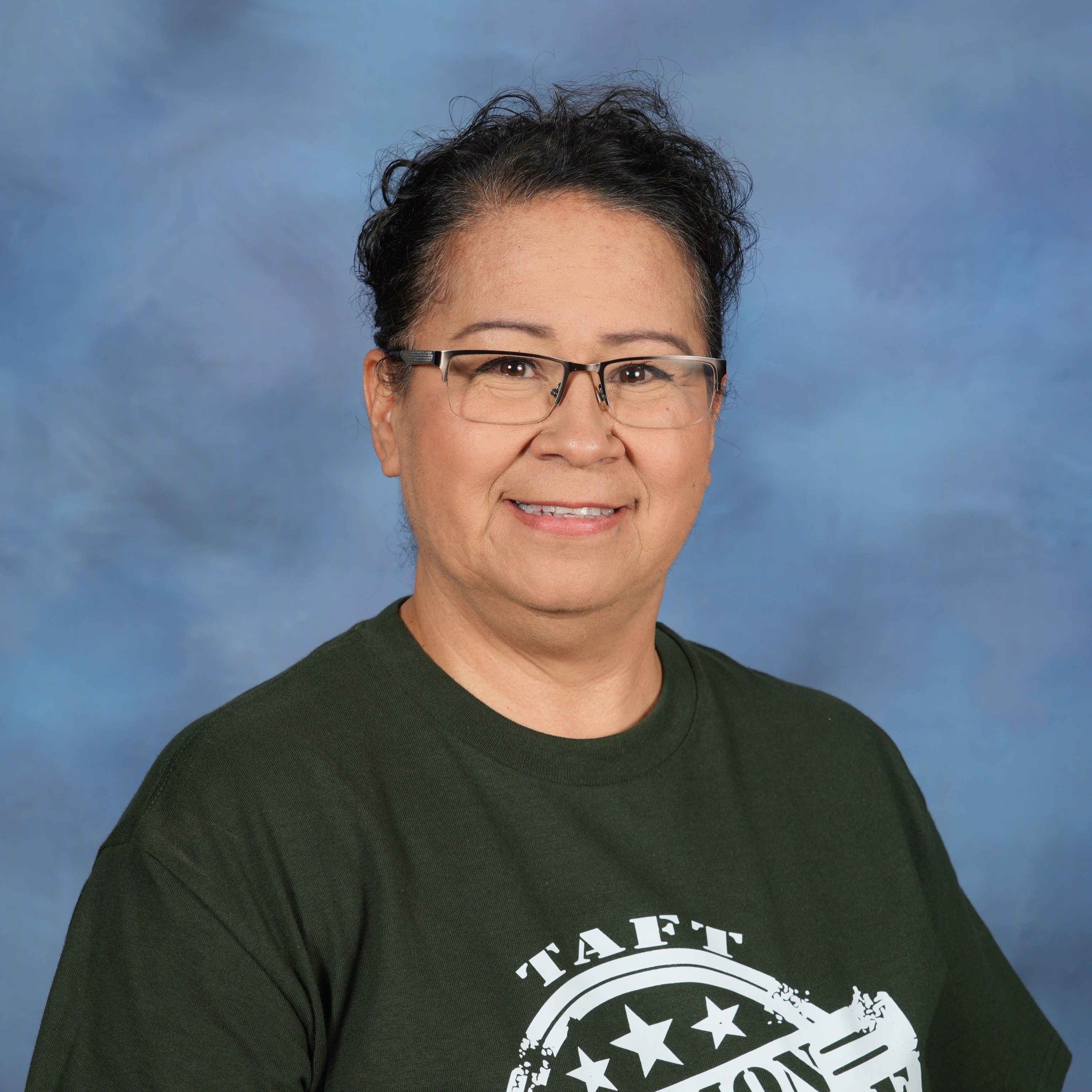 1. A woman wearing a green shirt and jeans stands confidently against a vibrant blue background.   2. A woman in a green shirt and jeans poses in front of a striking blue backdrop, showcasing her casual style.  3. A woman dressed in a green shirt and jeans stands before a bold blue background, exuding a relaxed demeanor.