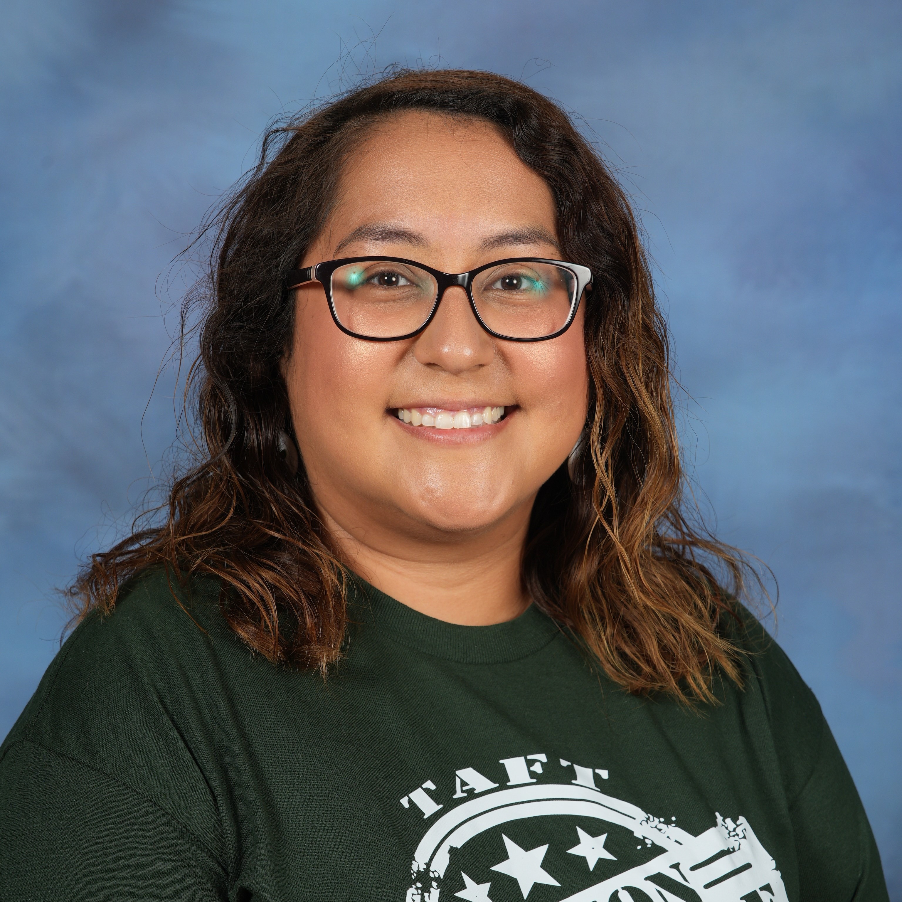A woman wearing a green shirt and glasses, smiling while standing against a neutral background.