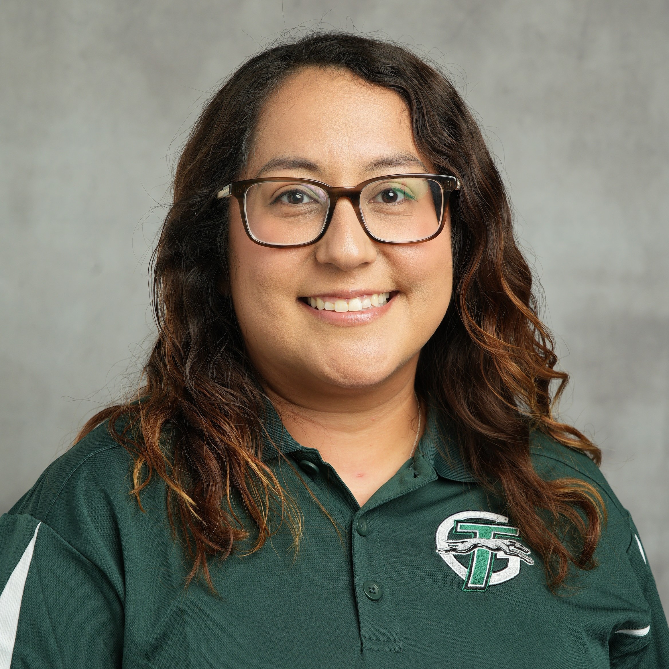 A woman wearing a green shirt and glasses, smiling while standing against a neutral background.