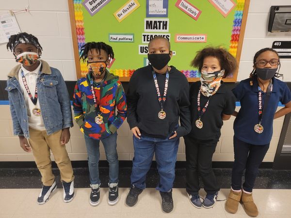 Five masked students with medals pose in front of a bulletin board, showcasing their achievements and school spirit.