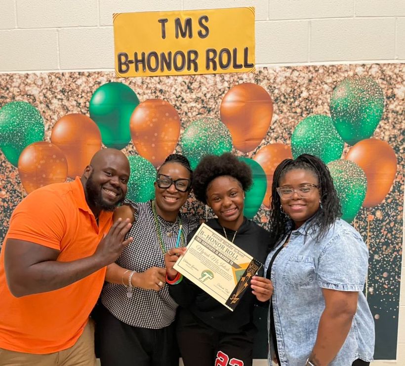 A group of three women and one man proudly display a plaque in front of a vibrant green wall.