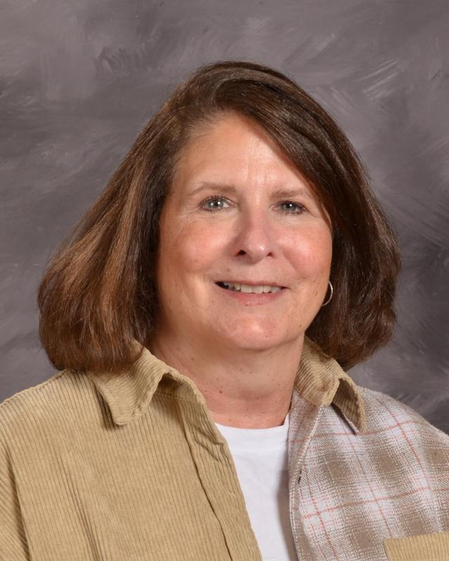 A woman wearing a tan shirt layered over a plaid shirt, standing confidently with a neutral background.