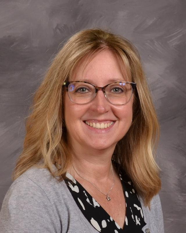 A woman wearing glasses and a gray cardigan smiles warmly at the camera.