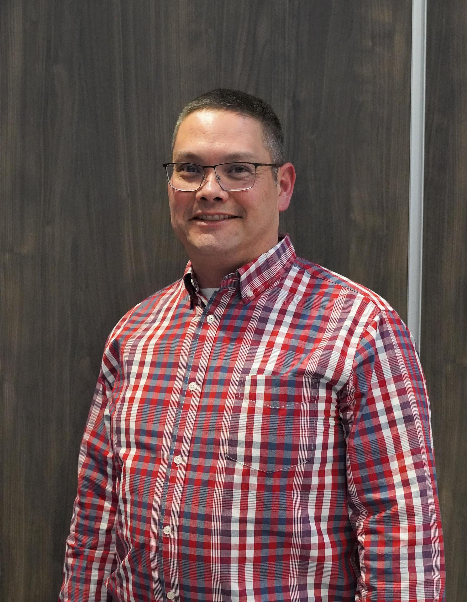 A man with glasses wearing a red and white checkered shirt, smiling and standing against a neutral background.
