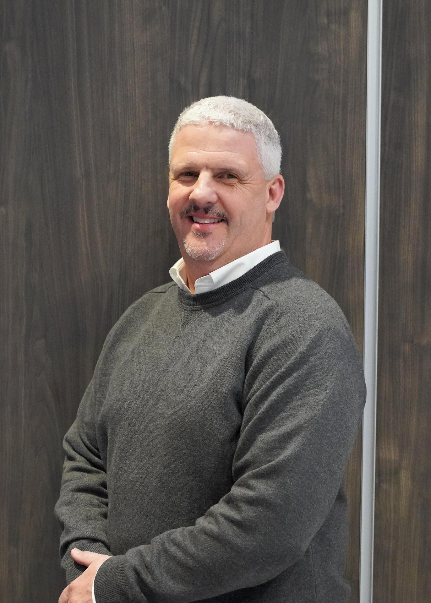 A man in a grey sweater stands confidently in front of a rustic wooden wall, showcasing a casual yet stylish appearance.