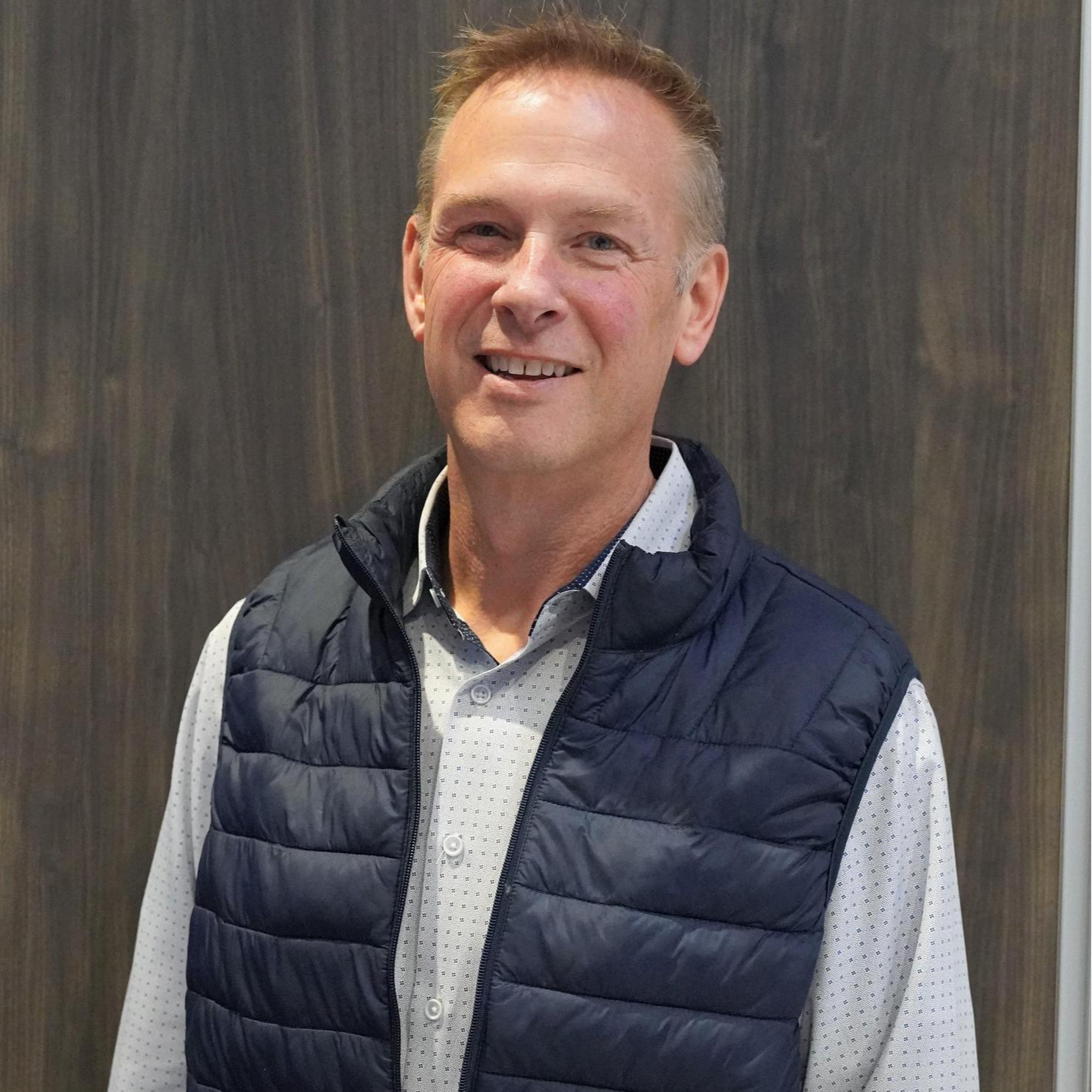 A man wearing a blue vest stands confidently in front of a rustic wooden wall.