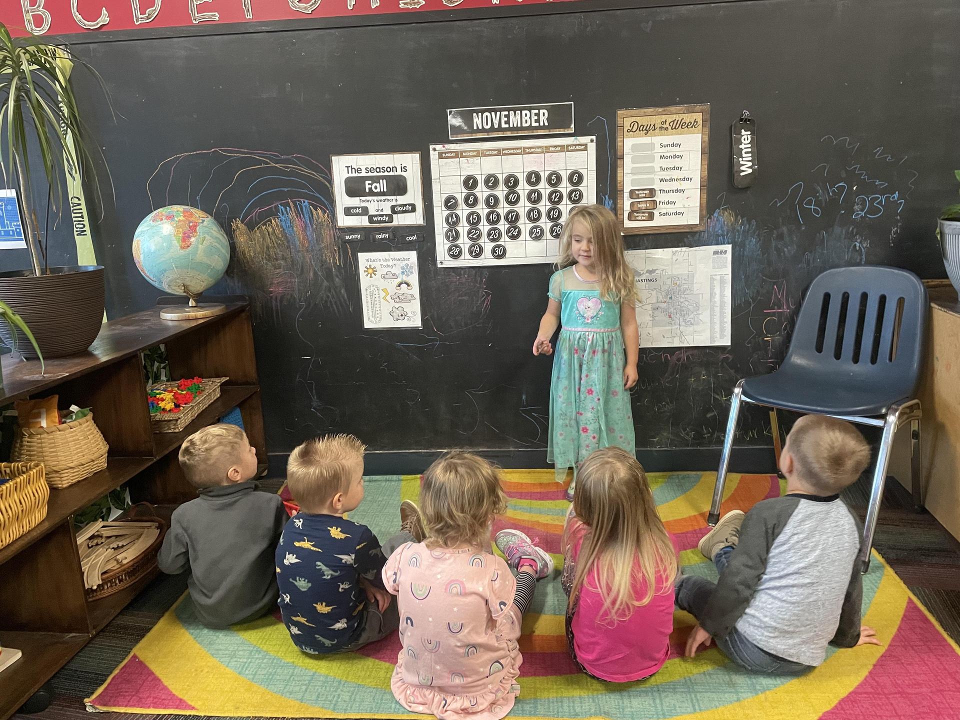 A group of children seated on the floor, engaged and focused, in front of a chalkboard filled with educational content.