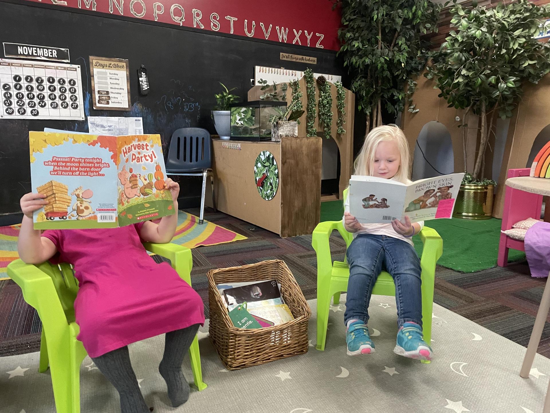 Two children seated in chairs, engrossed in reading books within a classroom setting.