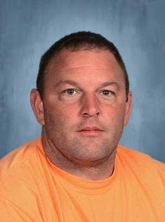 A man wearing an orange shirt poses confidently for a photograph, showcasing a cheerful demeanor.