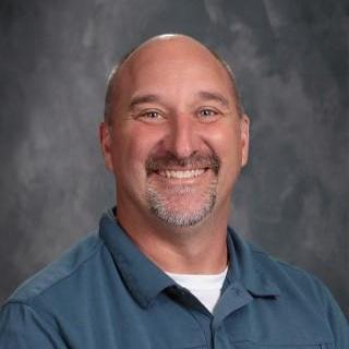 A man wearing a blue shirt smiles warmly, exuding a friendly and approachable demeanor.