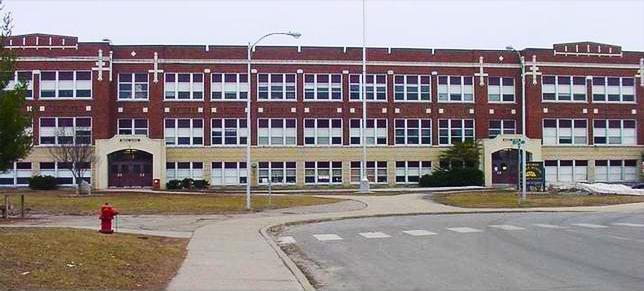 A historic brick school building featuring multiple large windows, a paved sidewalk, and manicured lawns, under an overcast sky.