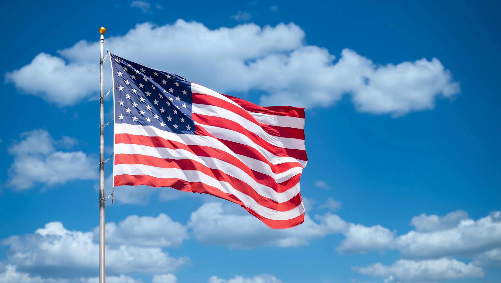 American flag waving against a clear blue sky with fluffy clouds.