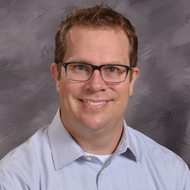 A man wearing glasses and a button down shirt smiles warmly at the camera.