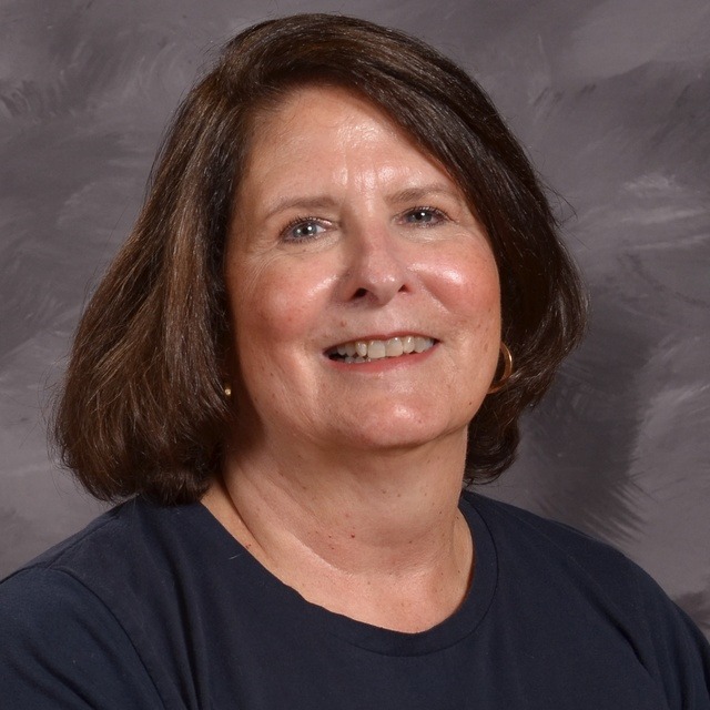 A woman wearing a tan shirt layered over a plaid shirt, standing confidently with a neutral background.