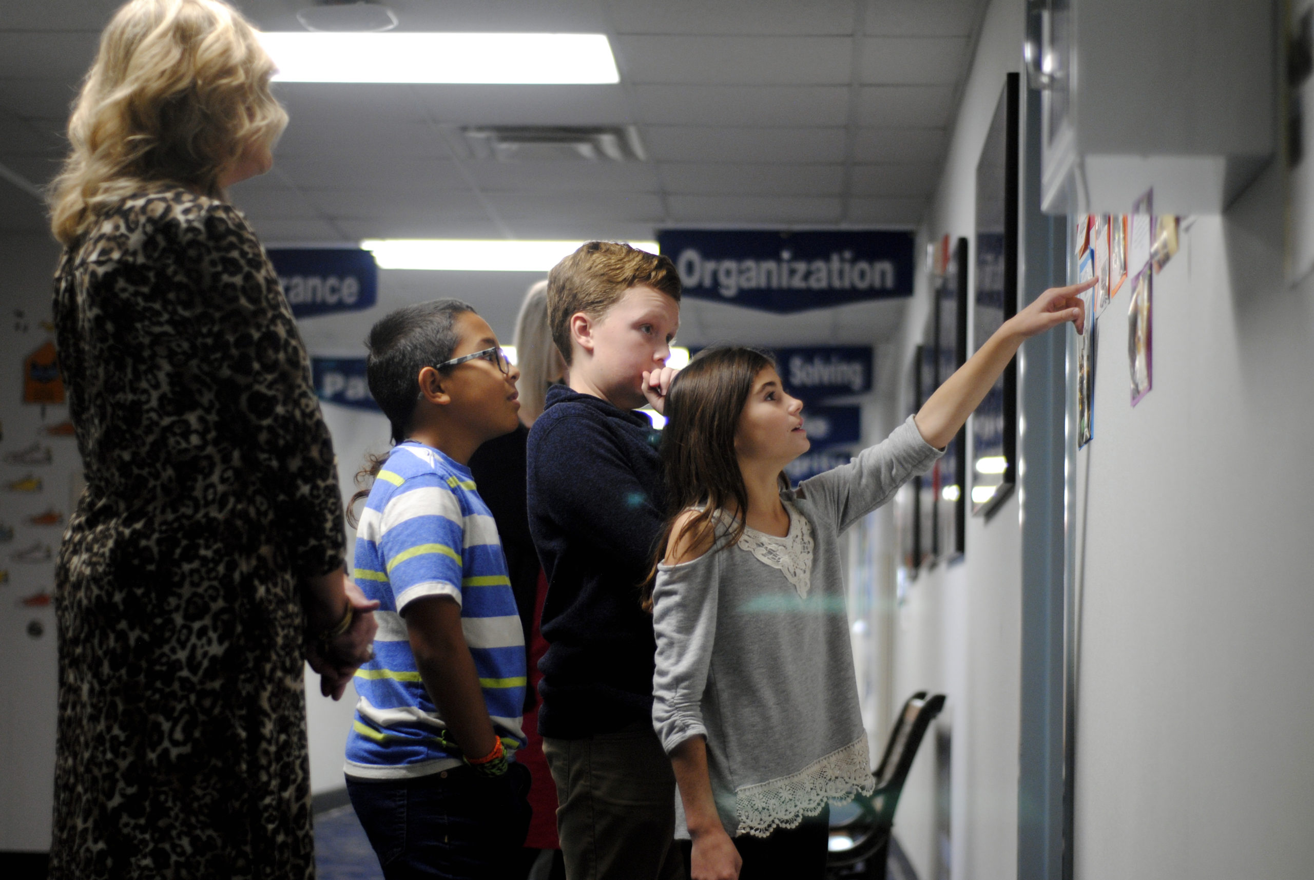 A woman stands beside a wall, pointing at a poster displayed prominently for viewers to see.