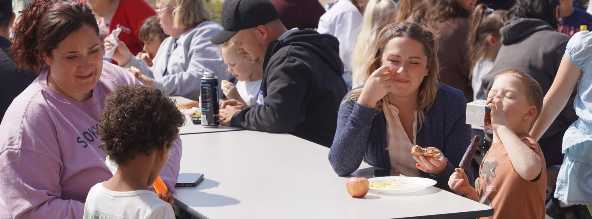 Parents and their children sit around a table eating at a cookout.