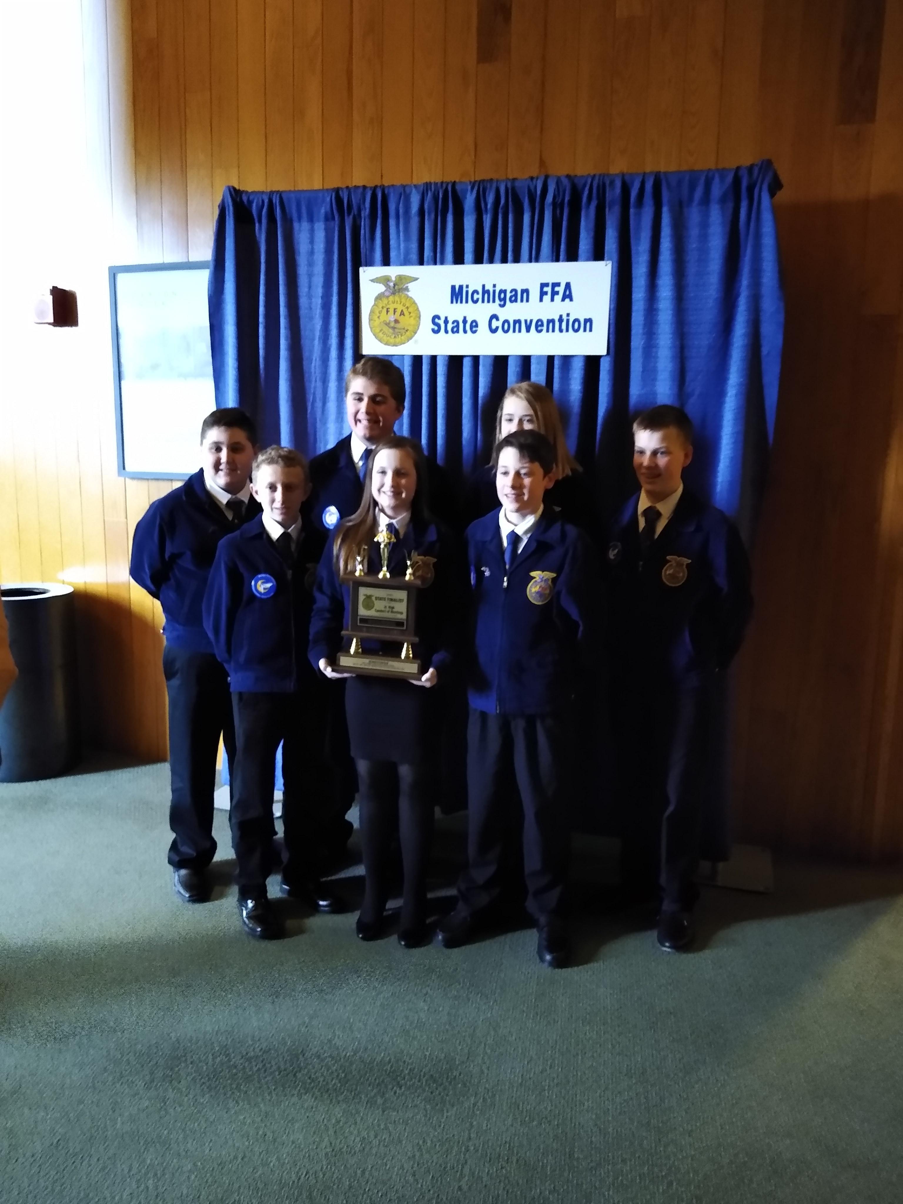 A group of students in FFA jackets pose proudly with a trophy in front of a Michigan FFA State Convention banner.