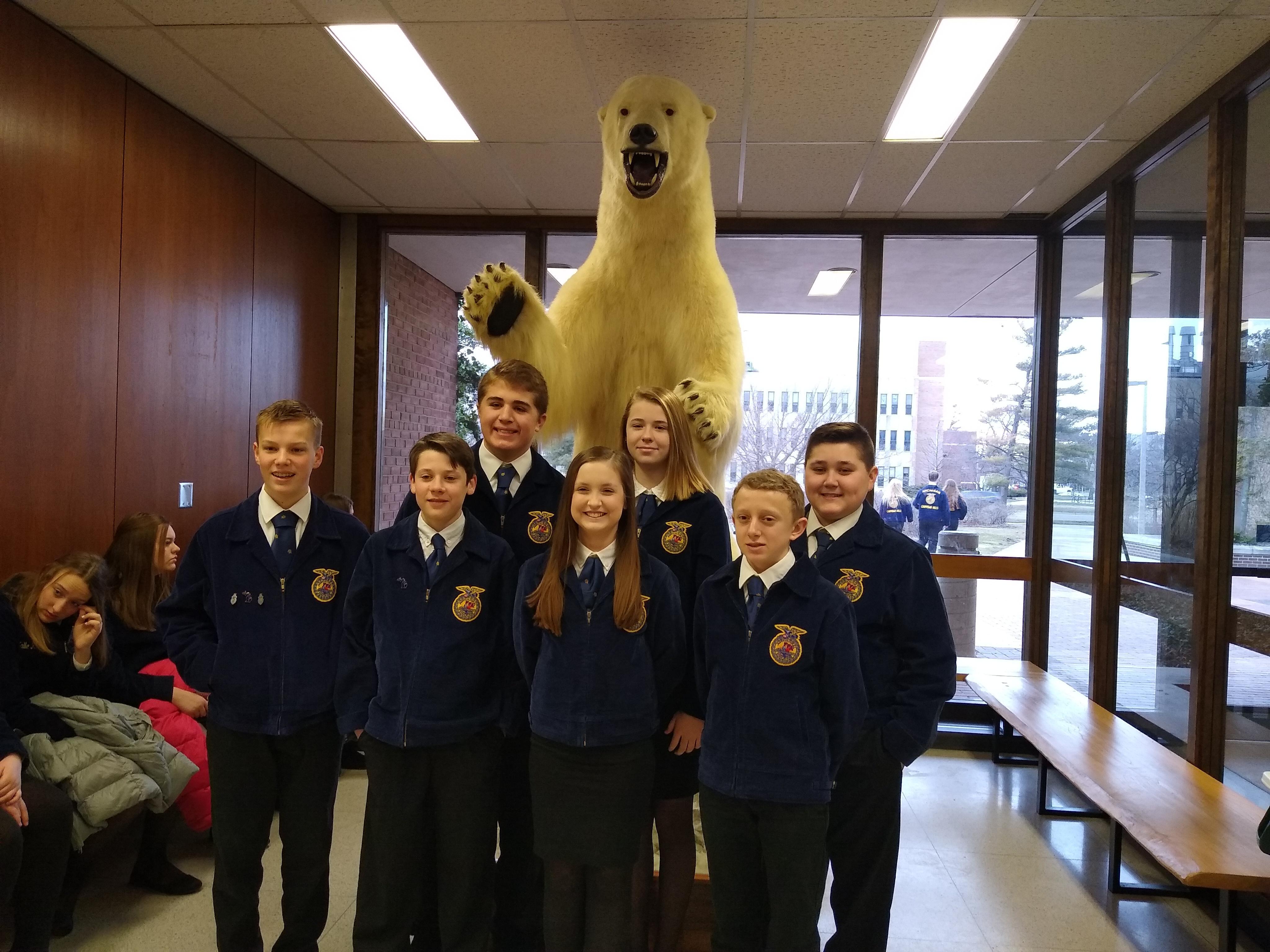 A group of students in blue FFA jackets stands in a school hallway, with a large mounted bear in the background. 
