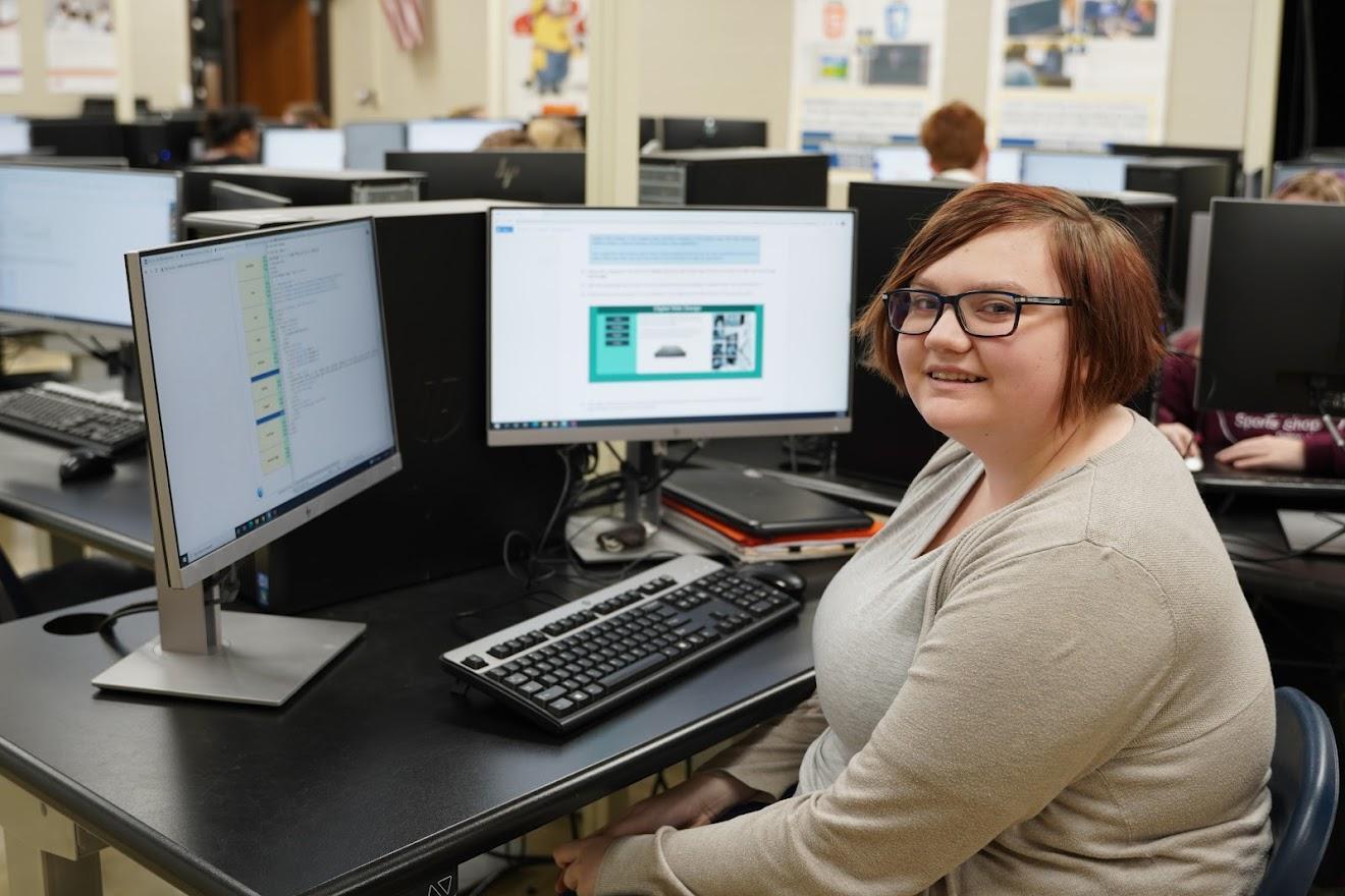 A student sits at a computer desk with multiple monitors displaying coding and design content in a classroom setting.