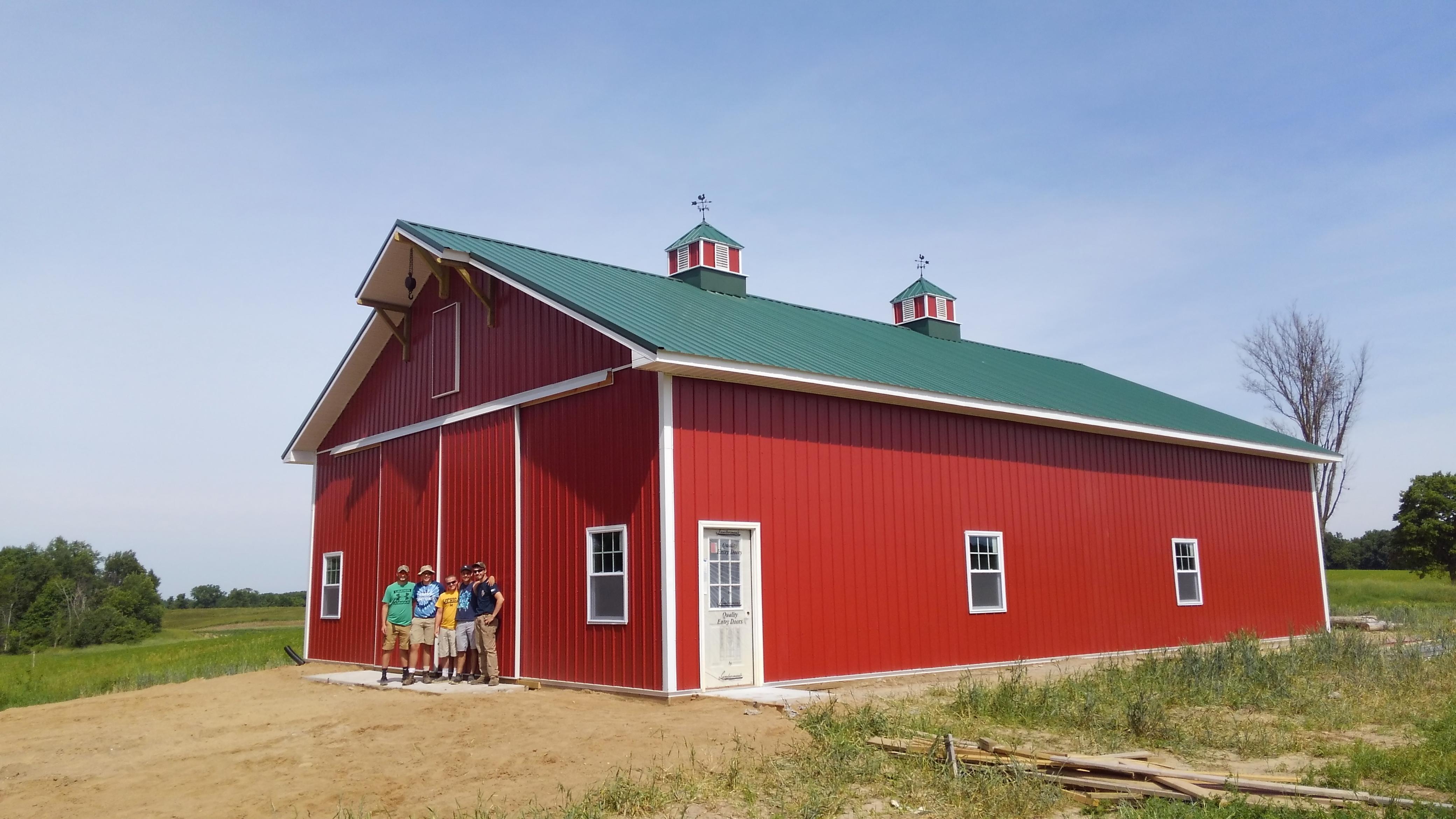A group of five people stand in front of a newly built red barn with a green roof and cupolas, surrounded by a grassy landscape.