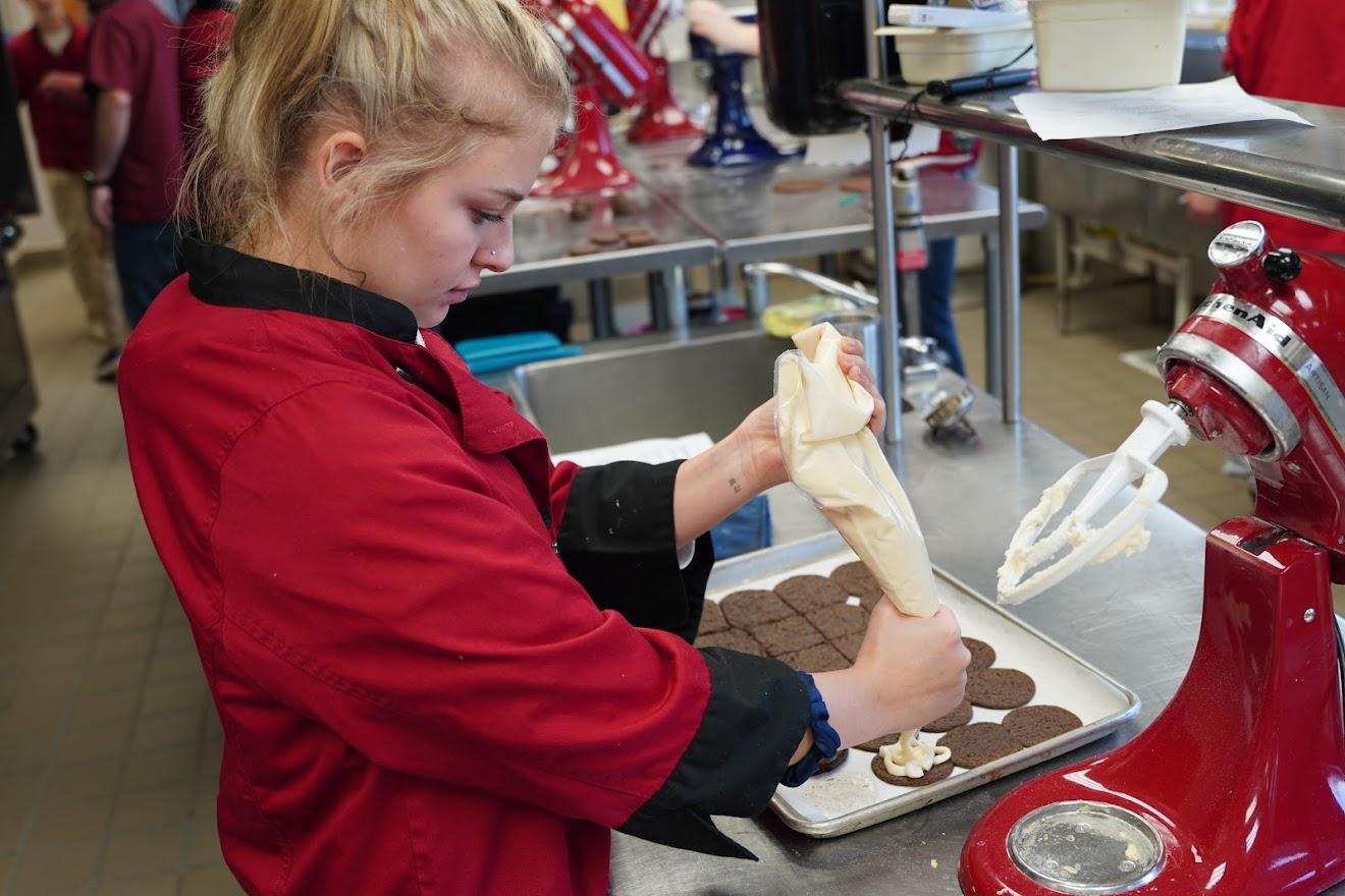 A person in a red apron pipes frosting onto chocolate cookies in a busy kitchen, with baking equipment and others in the background.