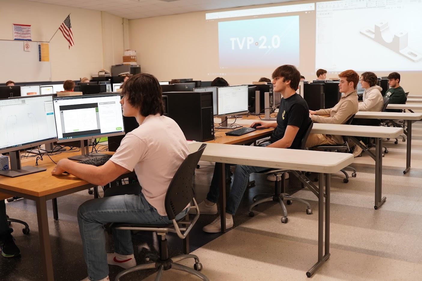 Students are seated at computers in a classroom, focused on screens displaying design software and instructional content.