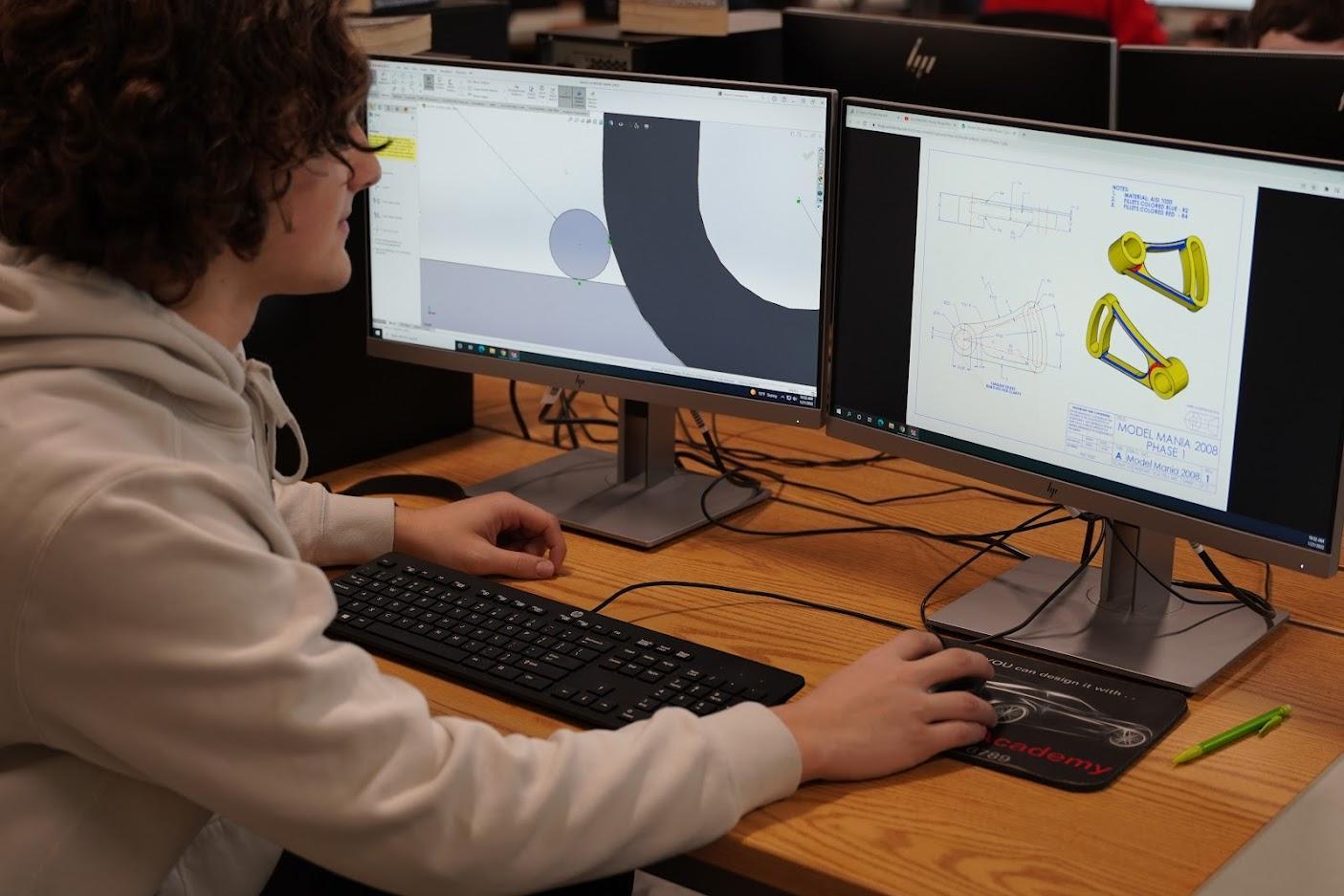 A student in a hoodie focuses on dual monitors displaying engineering designs and drawings in a classroom setting.