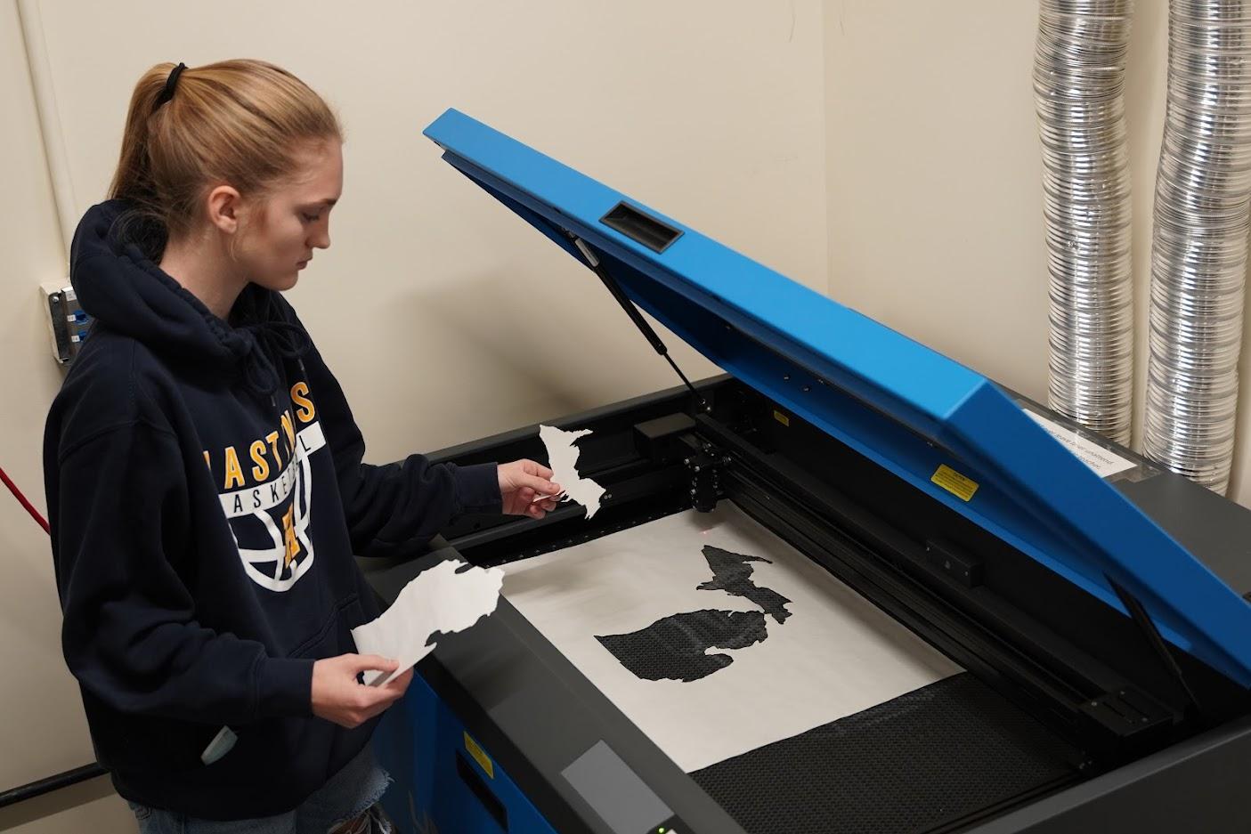 A person stands beside a laser cutter, holding cut paper shapes while the machine's lid is open, revealing a design underneath.