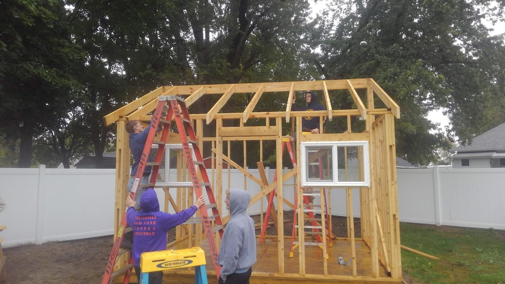 A construction site showing four individuals working on a wooden frame of a small building, with ladders and tools visible.