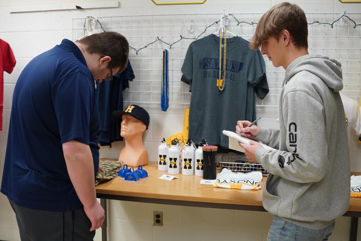 Two individuals stand beside a table filled with merchandise, including sports apparel, water bottles, and accessories.