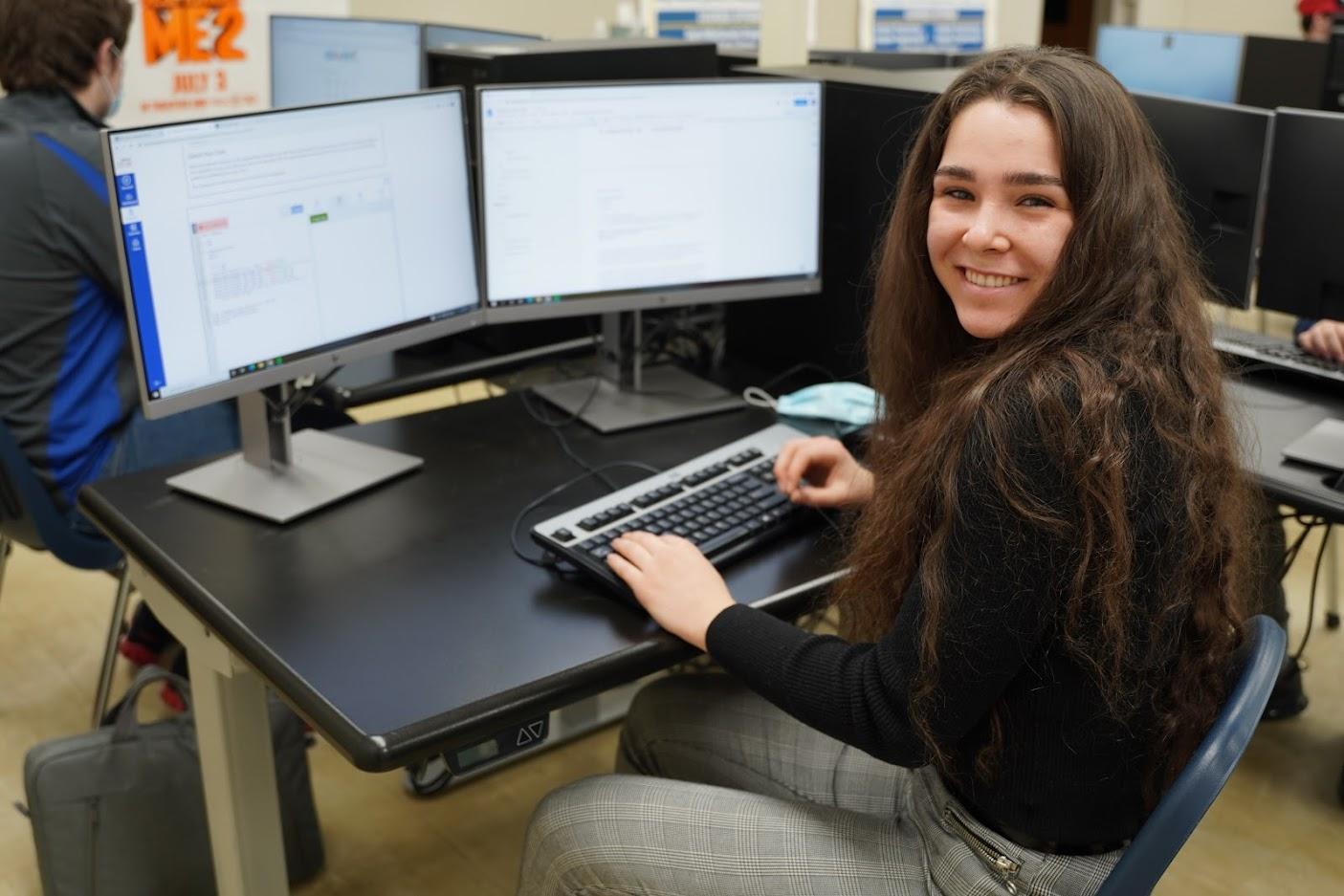 A student works on programming tasks at a dual-monitor setup in a classroom, with a laptop bag resting on the floor nearby.