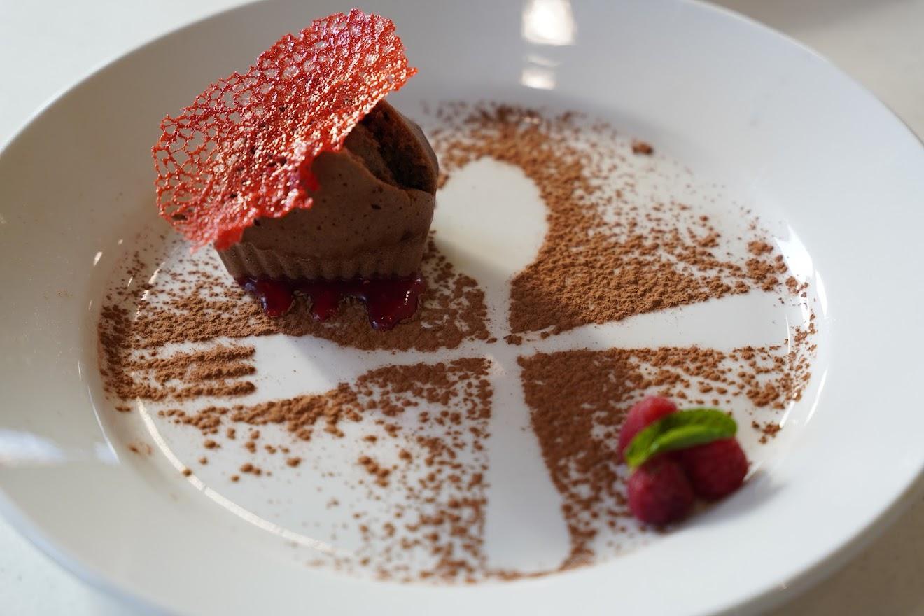 A gourmet chocolate dessert topped with a red sugar lace, surrounded by cocoa powder and fresh raspberries on a white plate.