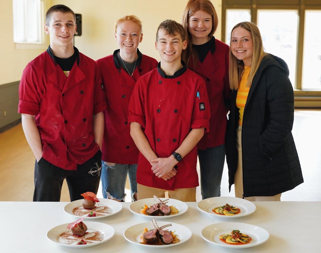 A group of young chefs in red jackets stands proudly behind a table displaying intricately plated gourmet dishes.
