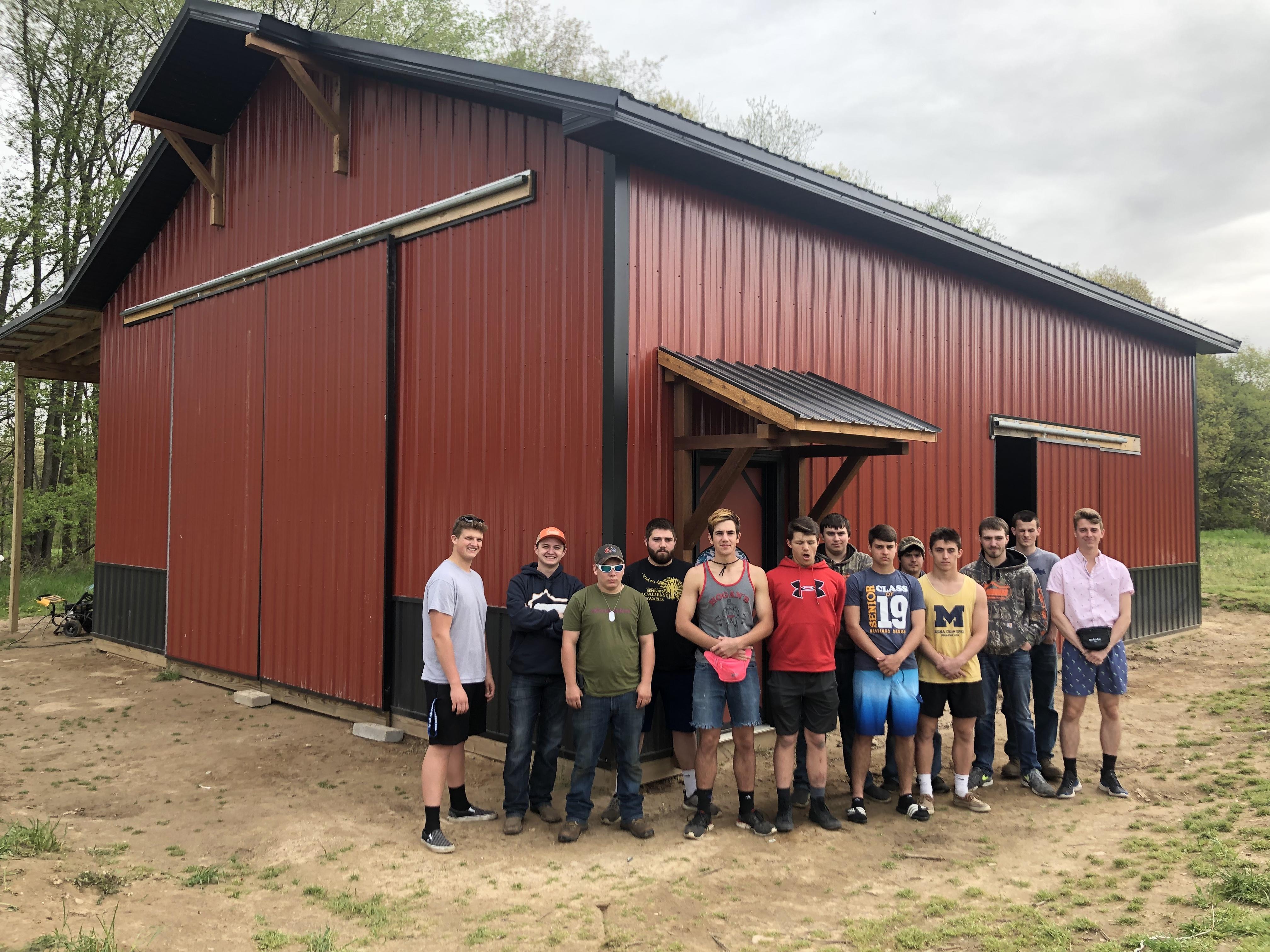 A group of individuals stands in front of a newly constructed red barn, surrounded by green trees and grass.