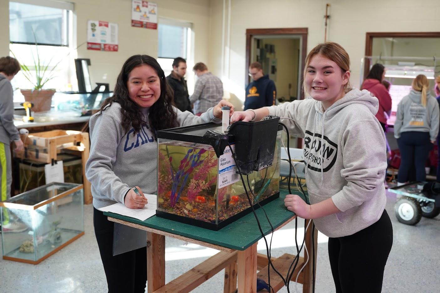 Two students are working together on an aquarium project, one taking notes while the other adjusts equipment on the tank.