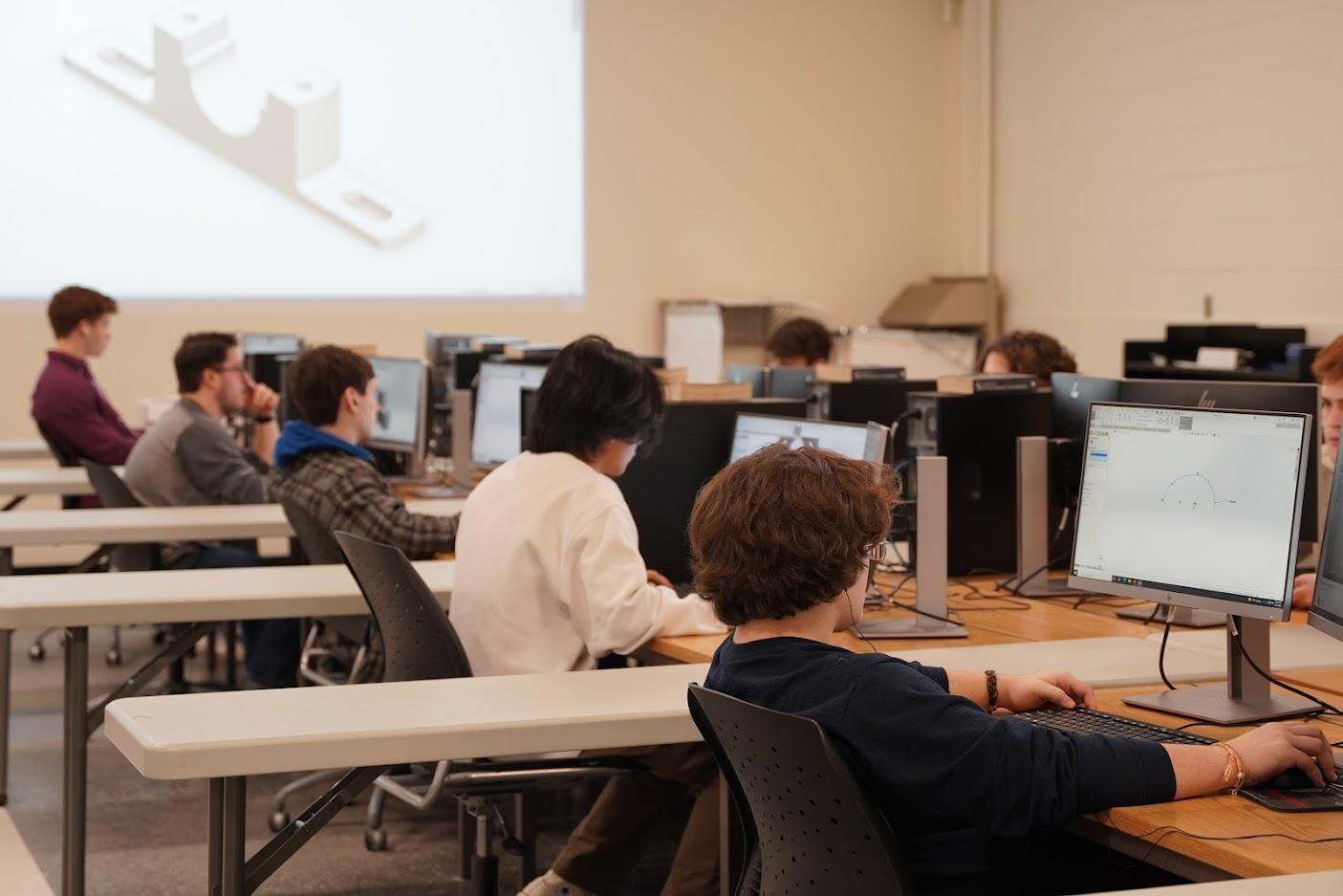 Students are seated at computer stations, working on design software in a classroom setting, with a 3D model projected behind them.