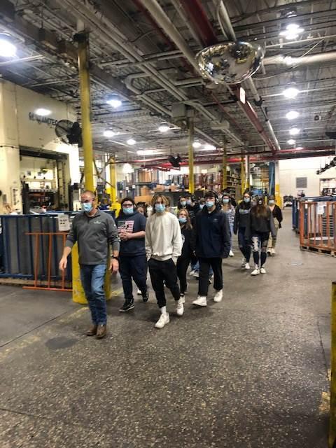 A group of masked individuals tours a large industrial facility, walking along a spacious aisle lined with equipment and machinery.