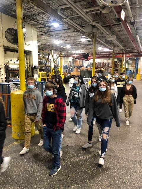 A group of masked students walks through a large industrial space, with metal structures and machinery in the background.
