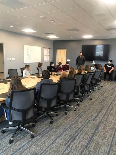 A group of students and adults in a conference room, engaging in a discussion, with a large screen and whiteboard in the background.