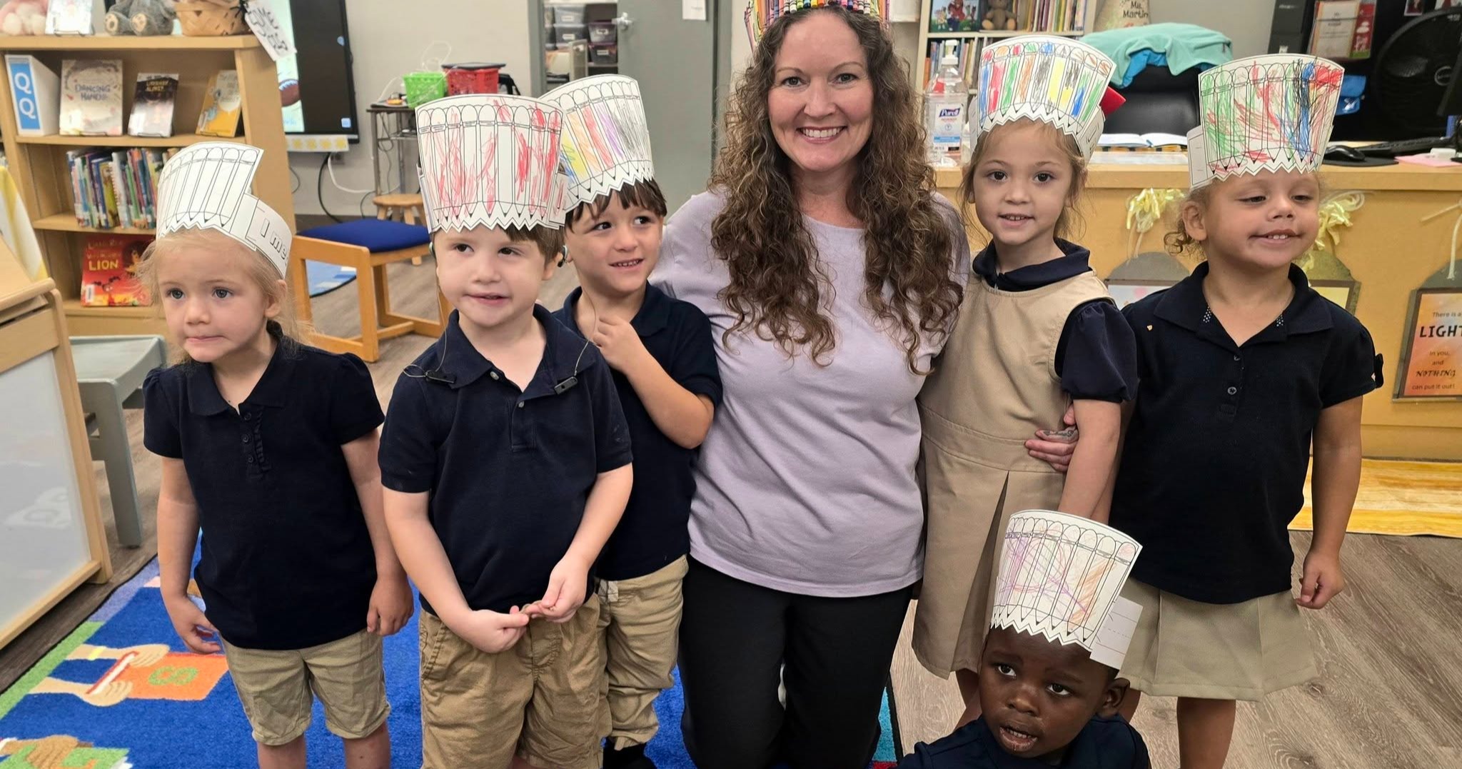 A woman stands with children in a classroom. The children wear cone hats. Behind them, a bookshelf is visible.