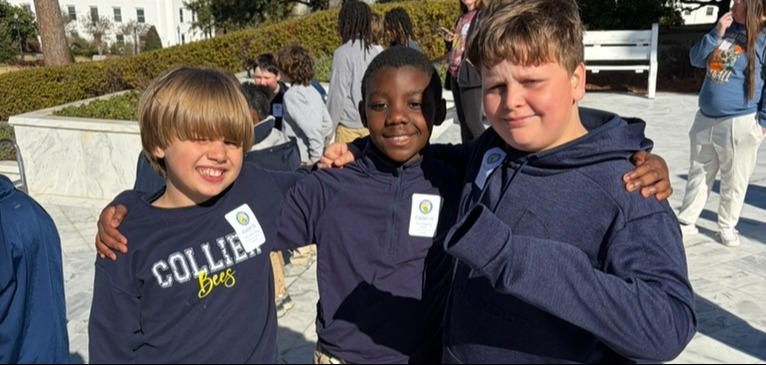 Three boys in matching blue hoodies and khaki pants pose for a photo with a white building and trees in the background.