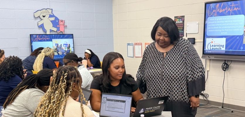 A group of people sit at tables in a classroom, using laptops. One person stands next to them.