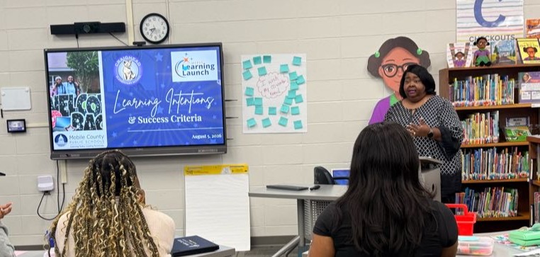 Classroom with a teacher at a podium, a screen on a wall, and students in chairs.