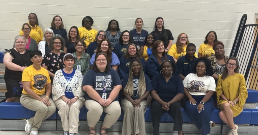 Group of people on bleachers in a school gym, most wearing blue shirts, some with yellow tops.
