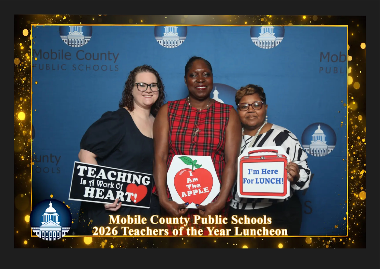 Three educators pose together in front of a “Mobile County Public Schools 2026 Teachers of the Year Luncheon” backdrop, each holding a celebratory sign, including messages such as “Teaching is a Work of Heart,” “I Am the Apple,” and “I’m Here for Lunch.”