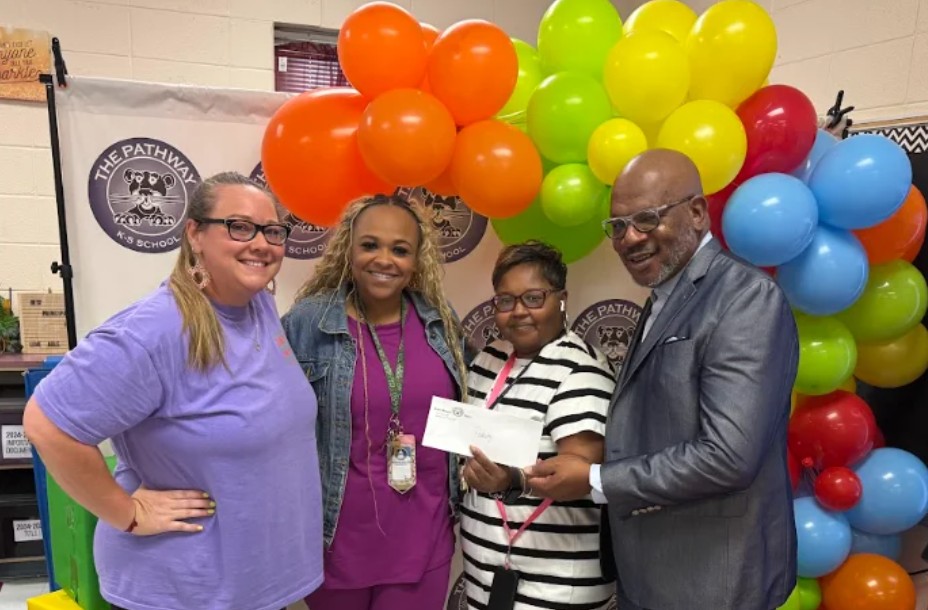 Four people pose for a photo in front of a large, multicolored balloon arch. One person holds a check.